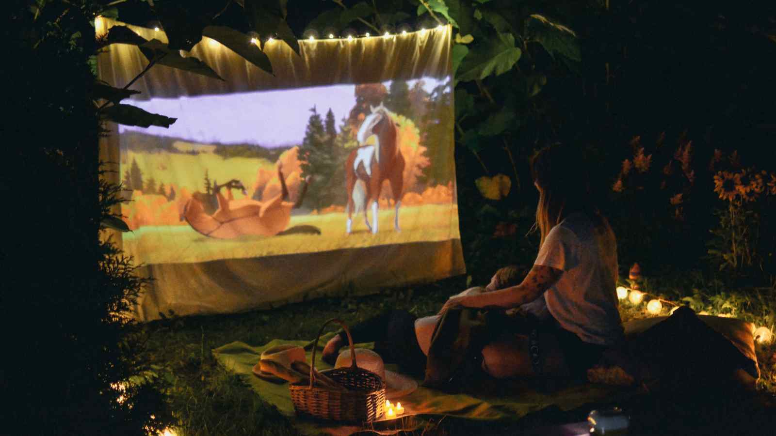 A person sits on a blanket outdoors at night, watching an animated movie projected onto a sheet, surrounded by string lights and a picnic basket.