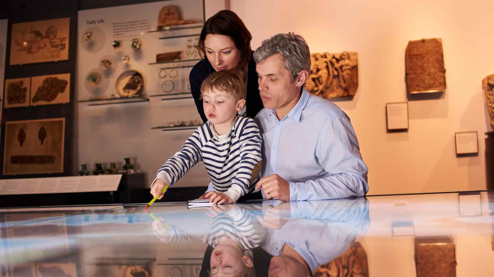 An adult man, woman, and young child interact with a large touchscreen display in a museum exhibit space.