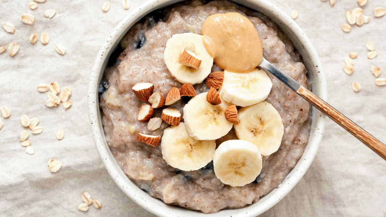Bowl of oatmeal topped with banana slices, chopped nuts, and a spoonful of peanut butter, with scattered oats on a beige surface.