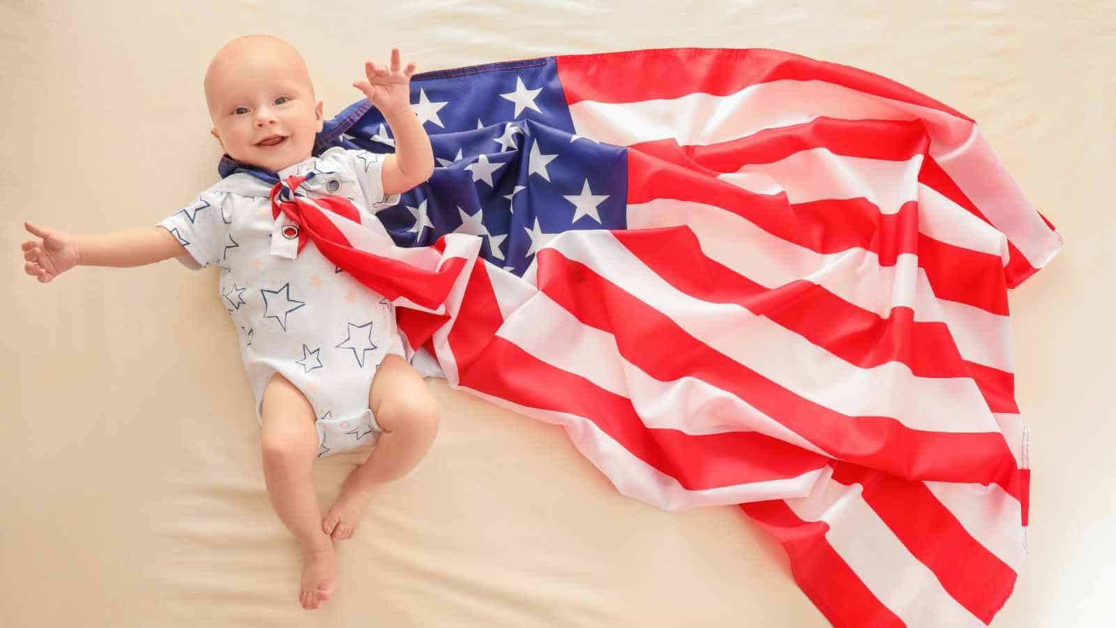 A baby in a star-patterned outfit lies on a light surface with an American flag draped beside him.