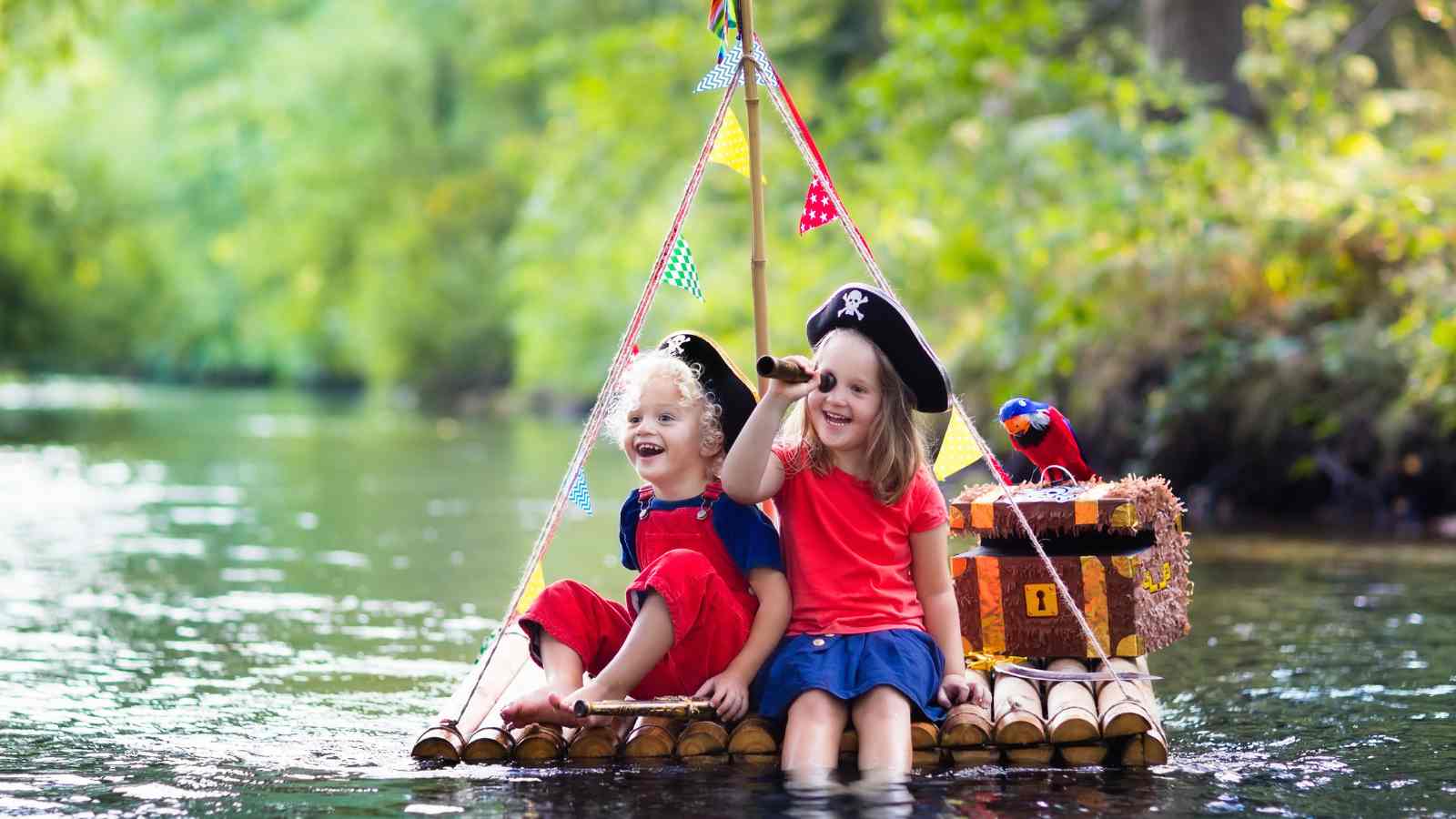 Two young children dressed as pirates sit on a homemade raft in a river, with a toy parrot and a treasure chest prop beside them.