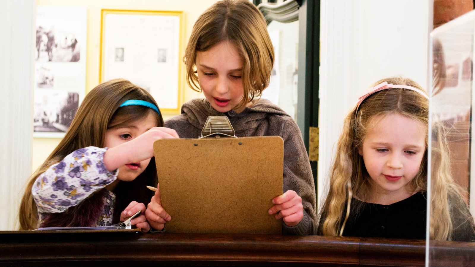 Three young girls stand together indoors, looking at a clipboard, appearing to work on an activity or project.