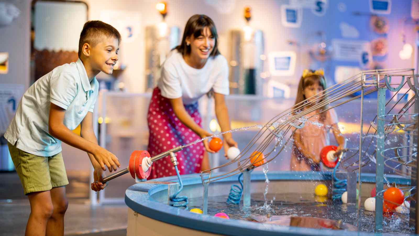 A boy, woman, and girl interact with a hands-on water exhibit, using tools to move balls along tracks in a brightly lit science center.