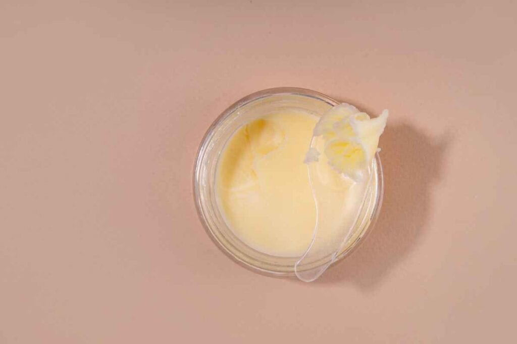 A glass jar filled with pale yellow cream sits on a neutral surface, with some of the cream on the rim.