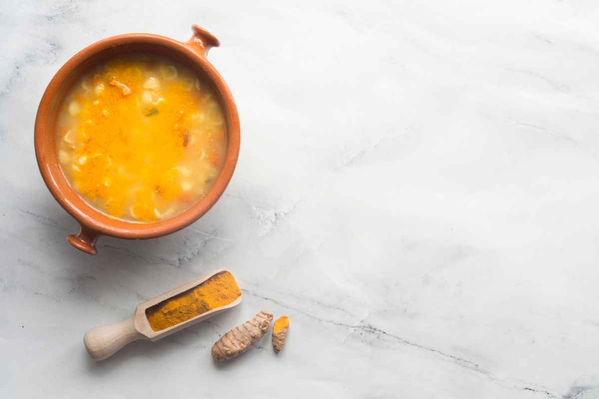 A bowl of yellow soup sits on a light surface next to a wooden scoop filled with turmeric powder and a piece of turmeric root.