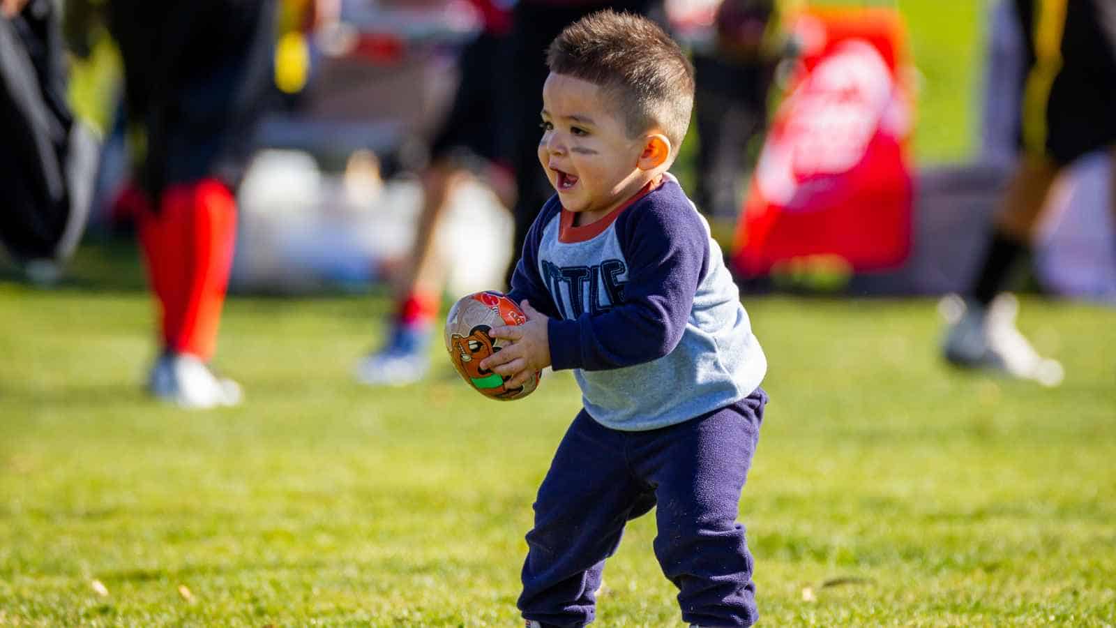 A young child holds a small football and runs on grass, with other people and sports equipment visible in the background.