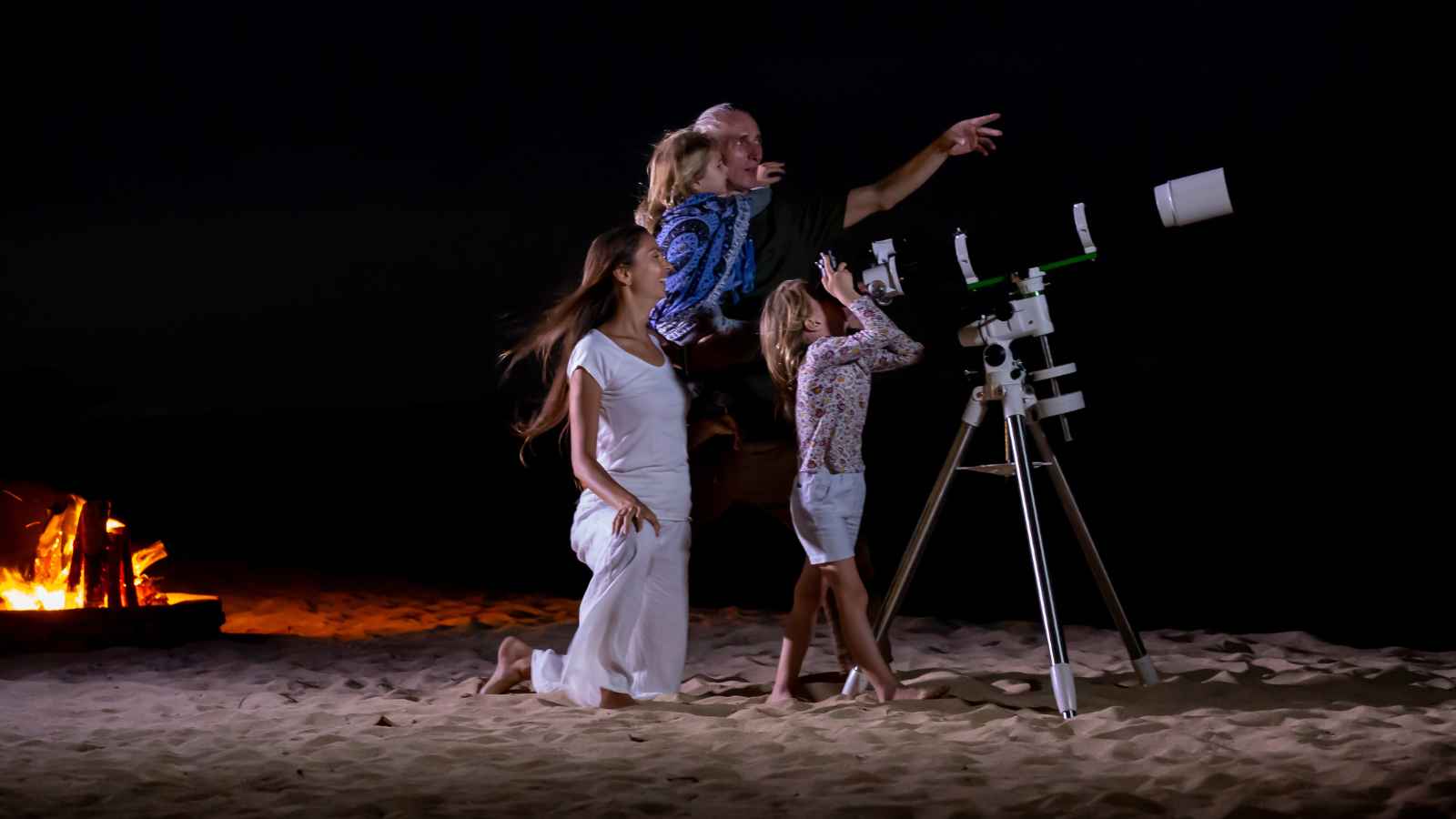 A family on a sandy beach at night looks through a telescope, with a campfire burning in the background.