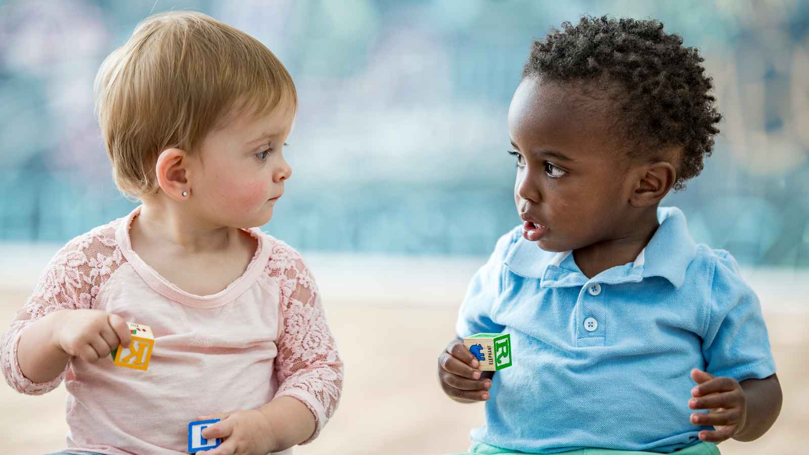 Two toddlers sit facing each other, holding lettered blocks.