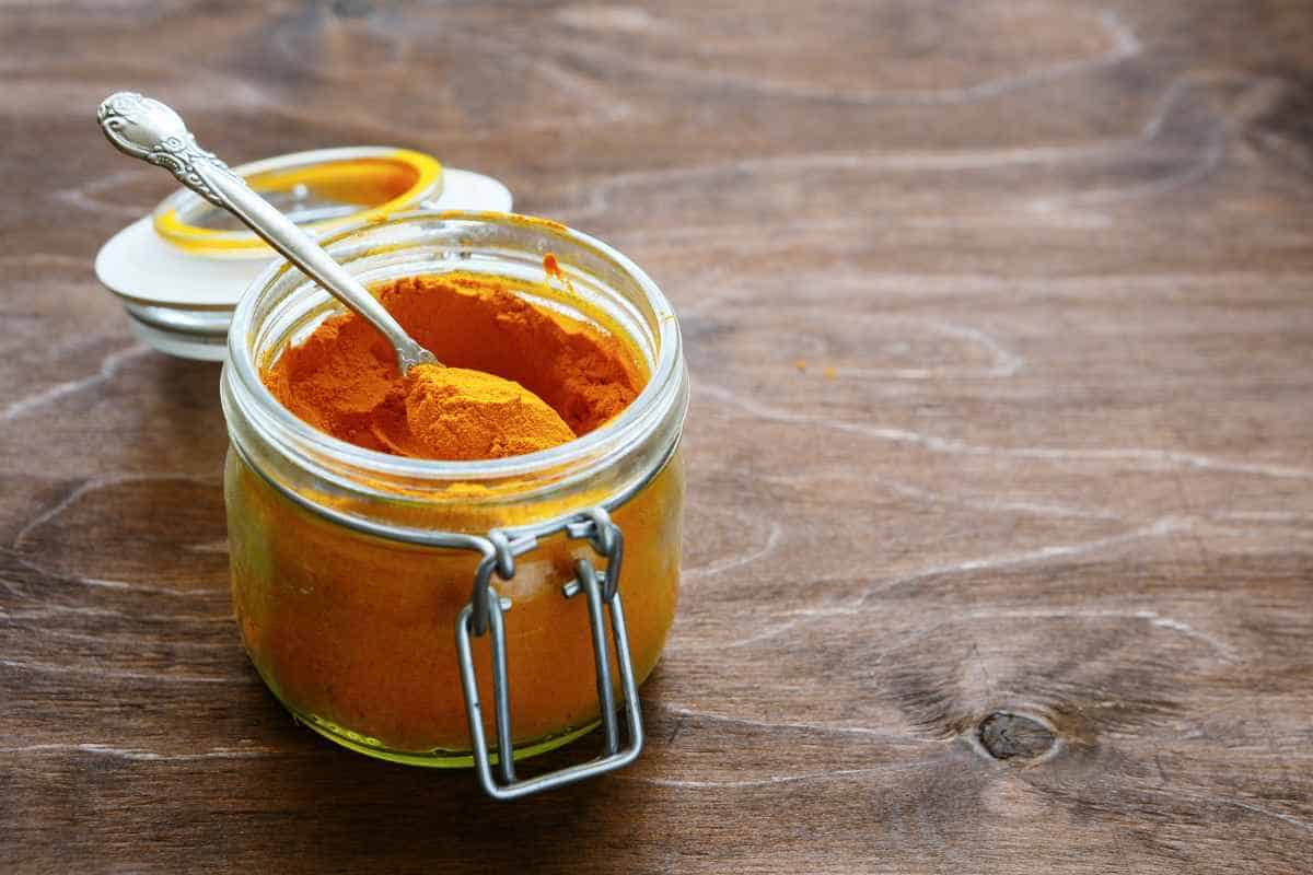 A glass jar filled with orange turmeric powder and a spoon, placed on a wooden surface.