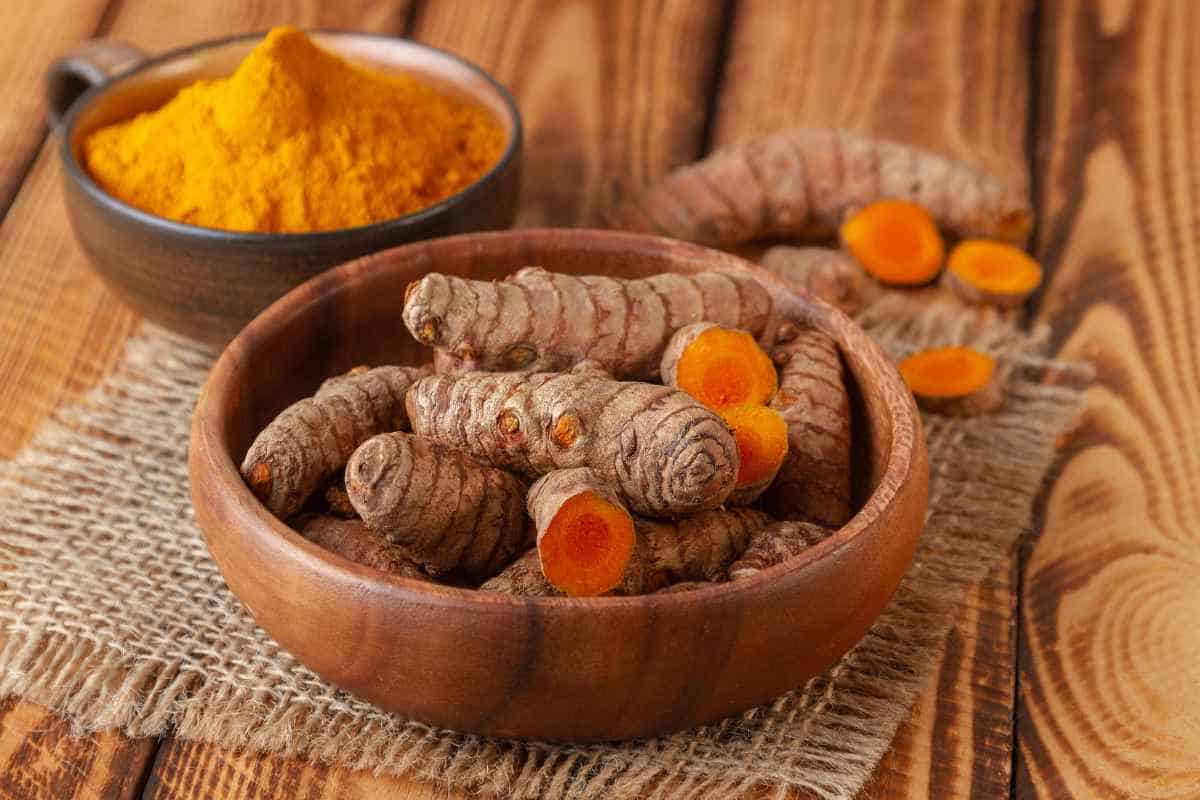 A wooden bowl filled with fresh turmeric roots, some cut to show the orange interior, with a bowl of turmeric powder in the background on a wooden surface.