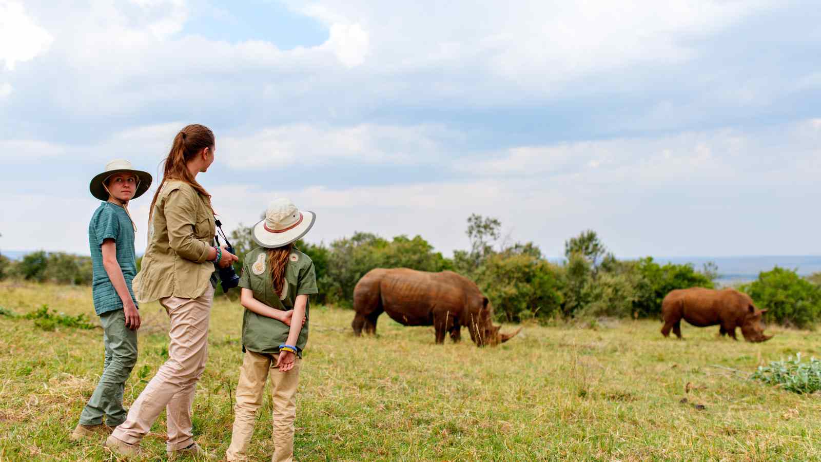 Three people in safari clothing observe three grazing rhinos in a grassy, open landscape with trees and cloudy sky in the background.