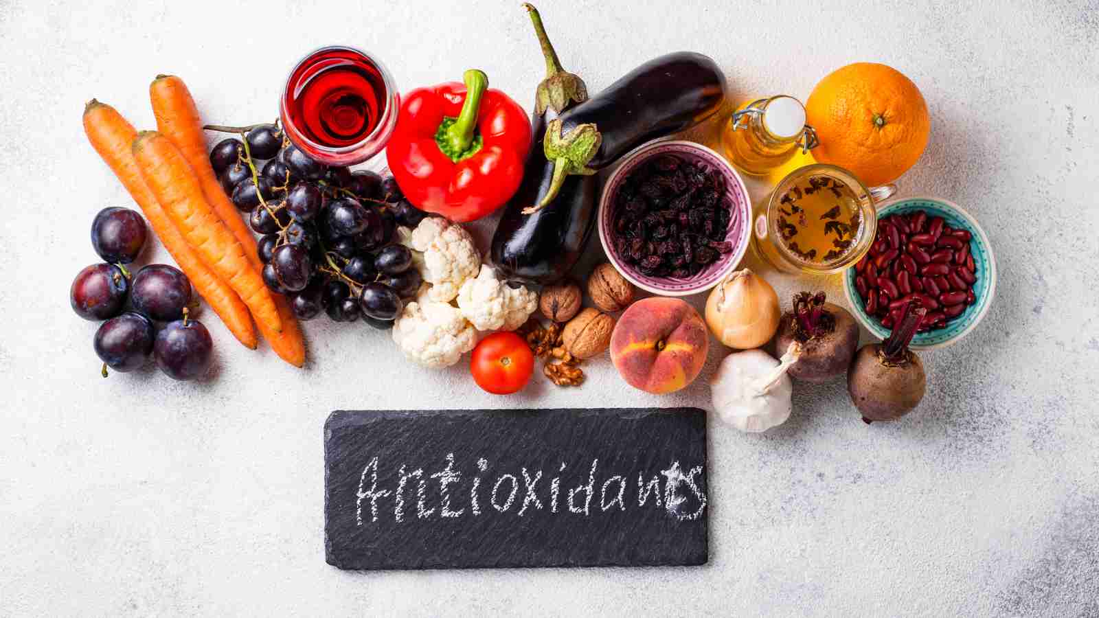 A variety of antioxidant-rich foods, including fruits, vegetables, nuts, and seeds, are arranged around a small chalkboard with the word "Antioxidants" written on it.