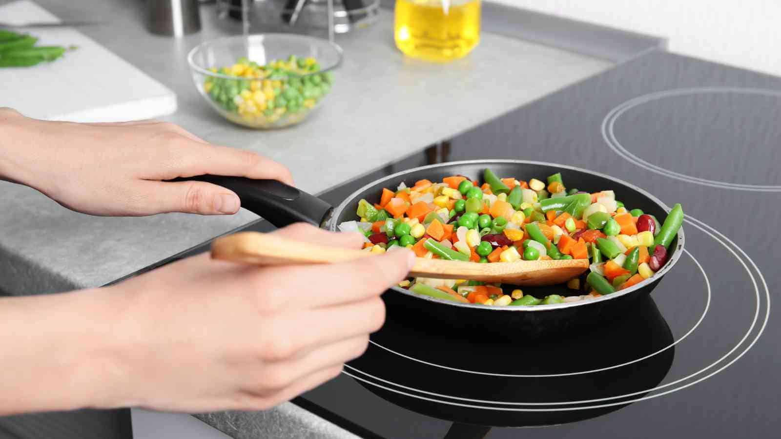 A person cooks a mix of chopped vegetables in a frying pan on an electric stovetop, stirring with a wooden spatula.