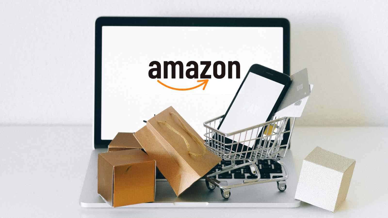 A laptop displaying the Amazon logo is in the background, with a miniature shopping cart, smartphone, credit card, and shopping bags placed in front.