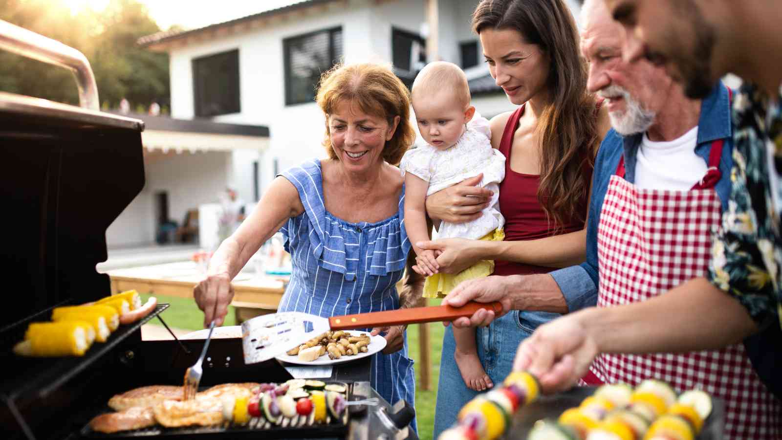 A group of people, including a woman holding a baby, gather around an outdoor grill cooking food in a backyard setting.