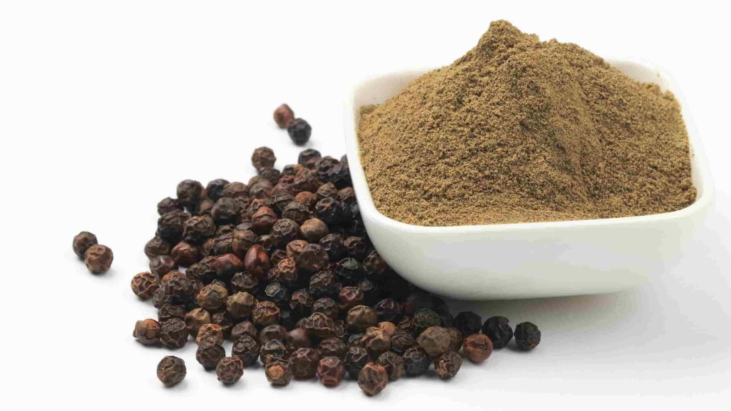 A pile of whole black peppercorns next to a white bowl filled with ground black pepper on a white background.