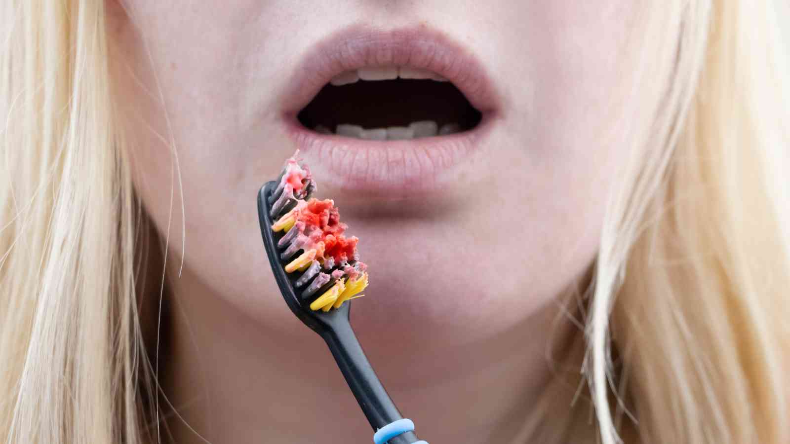 A close-up of a person with an open mouth holding a toothbrush covered in red substance resembling blood.