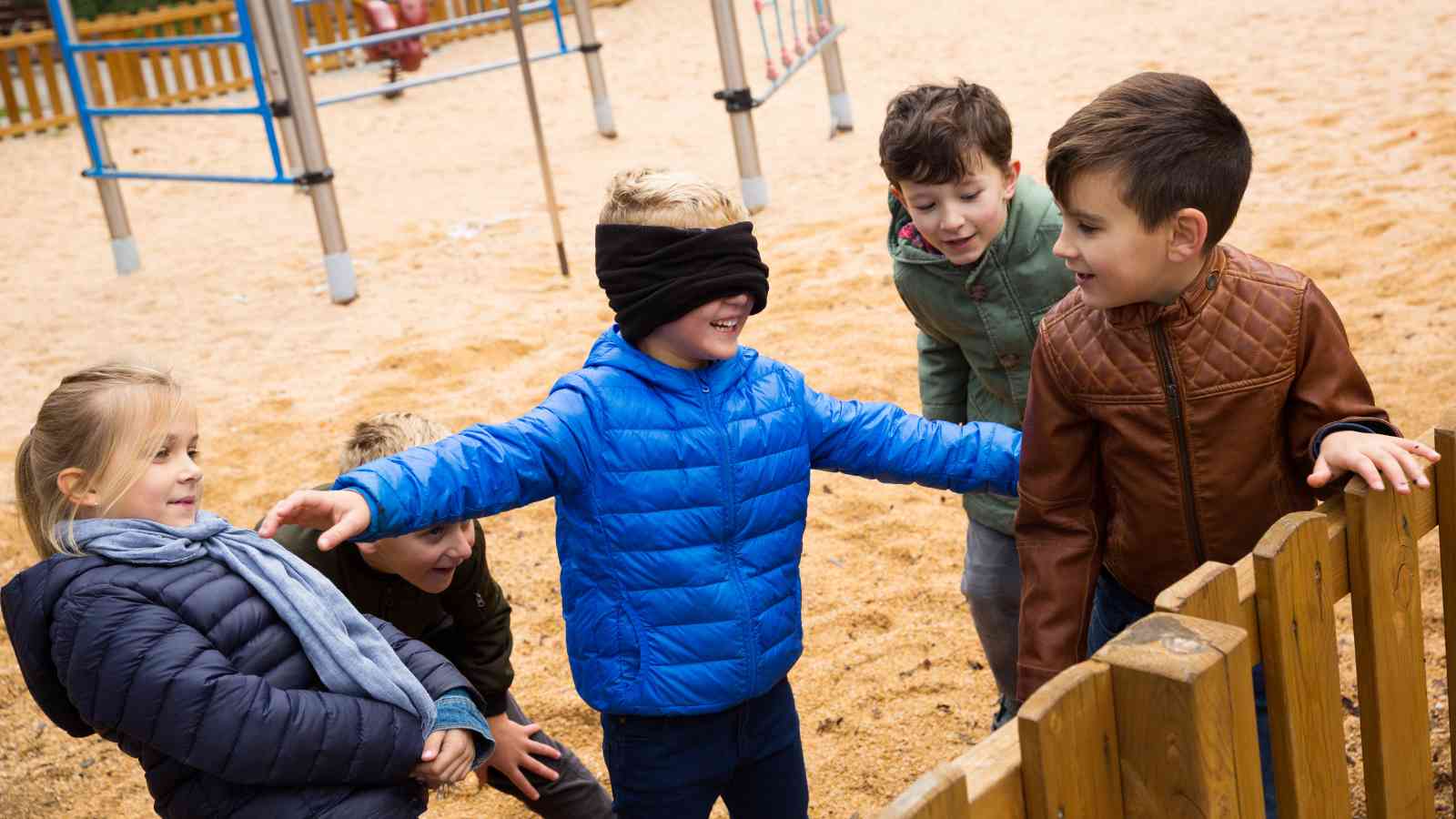 Five children play outside on a playground; one child is blindfolded and reaching out while the others watch and smile.