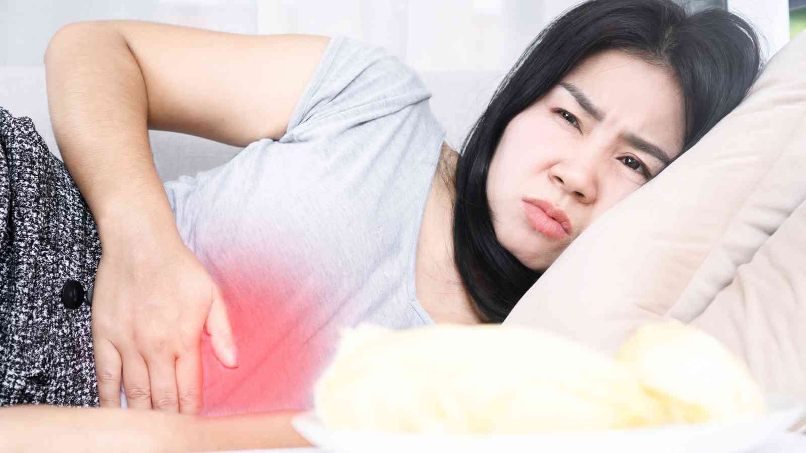 Woman lying on a couch, holding her stomach with a pained expression, suggesting abdominal discomfort or pain.