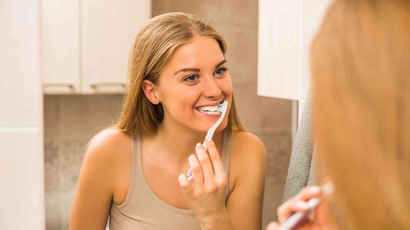 A woman with long hair is brushing her teeth in front of a bathroom mirror, wearing a beige tank top and smiling.