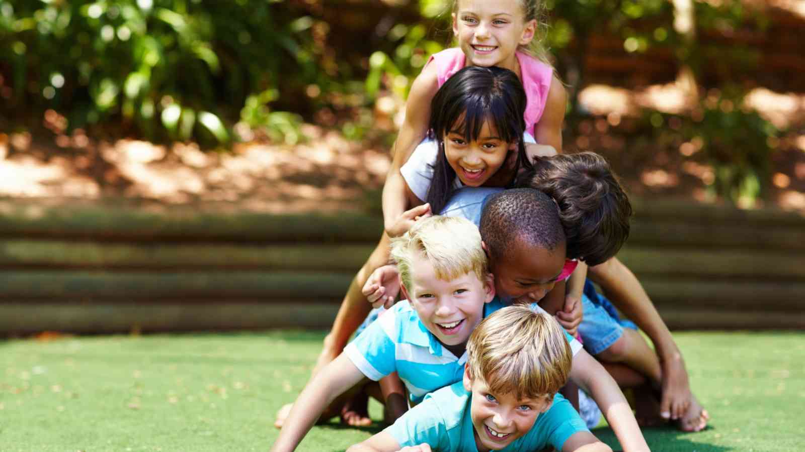 Six children form a playful human pyramid on grass outdoors, smiling and enjoying a sunny day. Trees and wooden barriers are visible in the background.