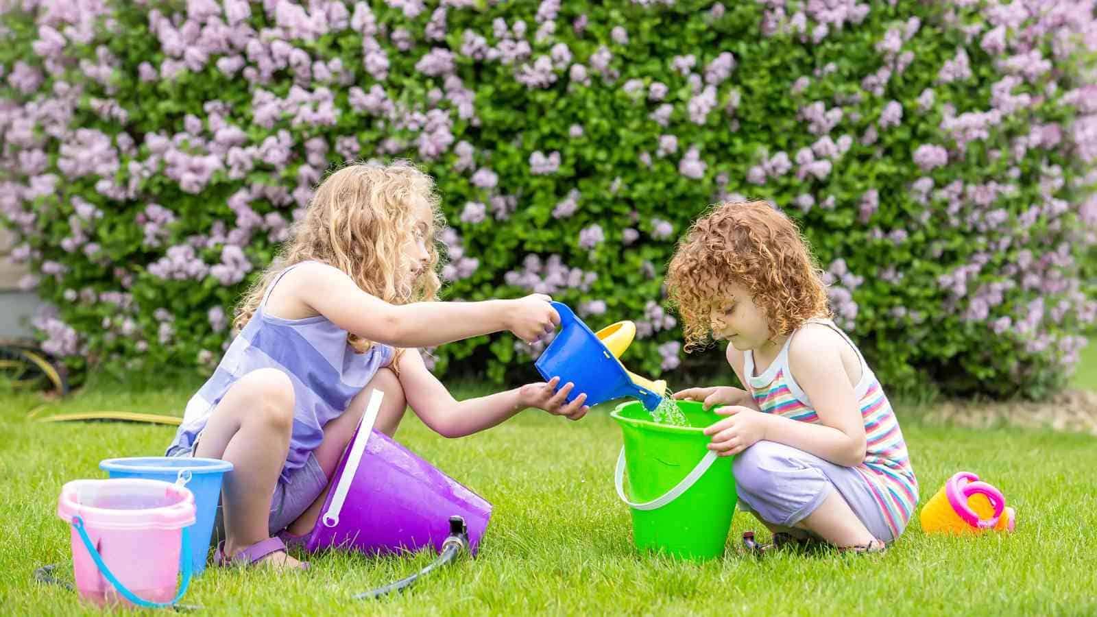 Two young children sit on grass, pouring water from a blue bucket into a green bucket, with colorful buckets and blooming bushes in the background.