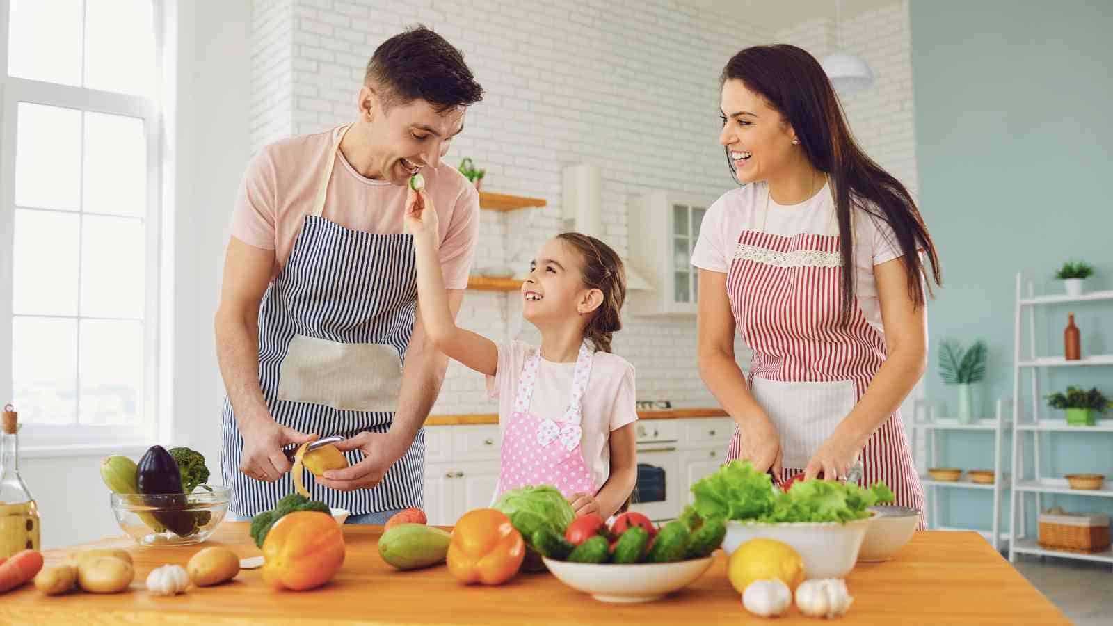 A man, woman, and young girl in aprons prepare vegetables together at a kitchen counter, smiling and enjoying the activity.