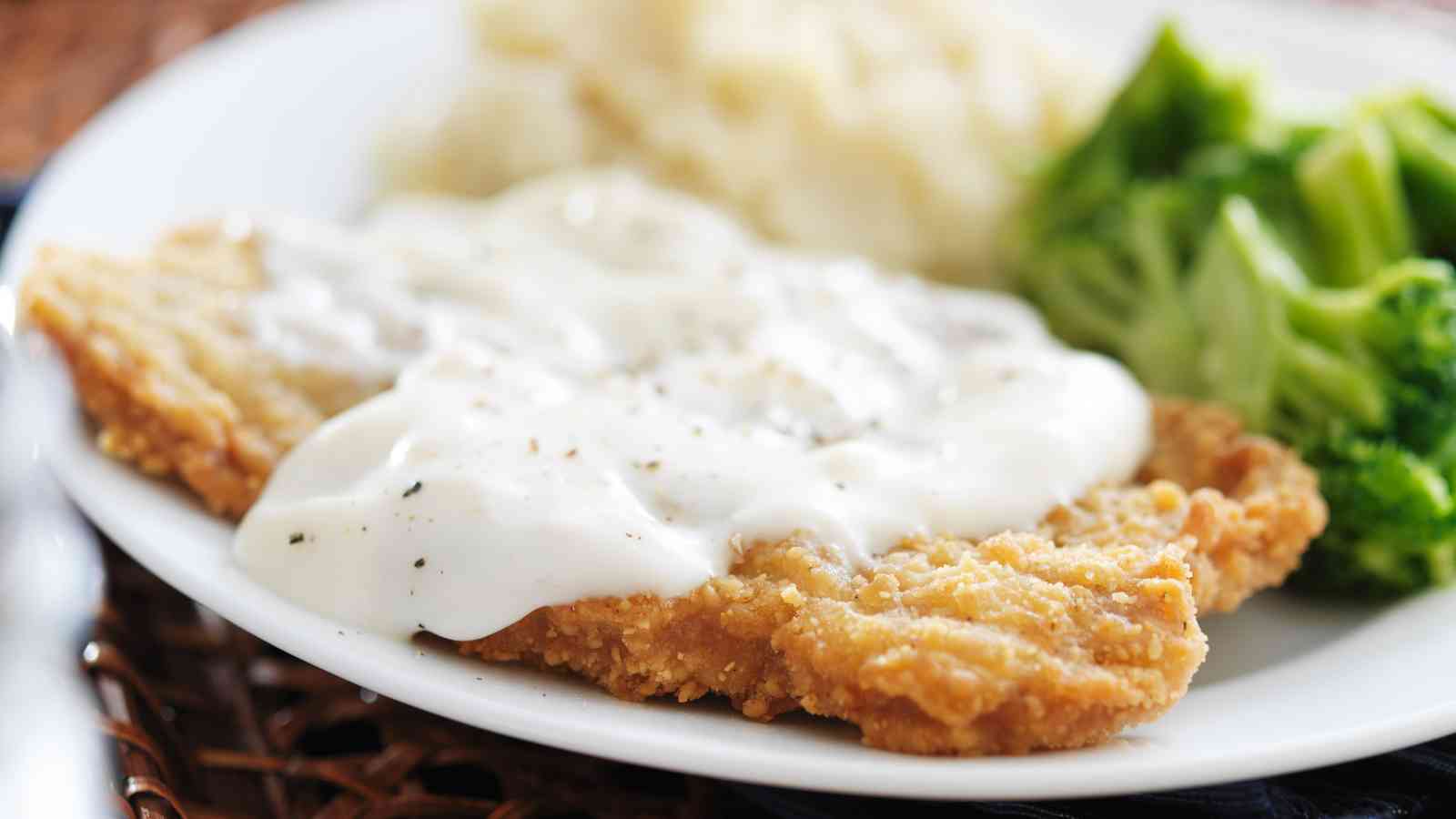 A plate of breaded fried chicken covered in white gravy, served with mashed potatoes and steamed broccoli.
