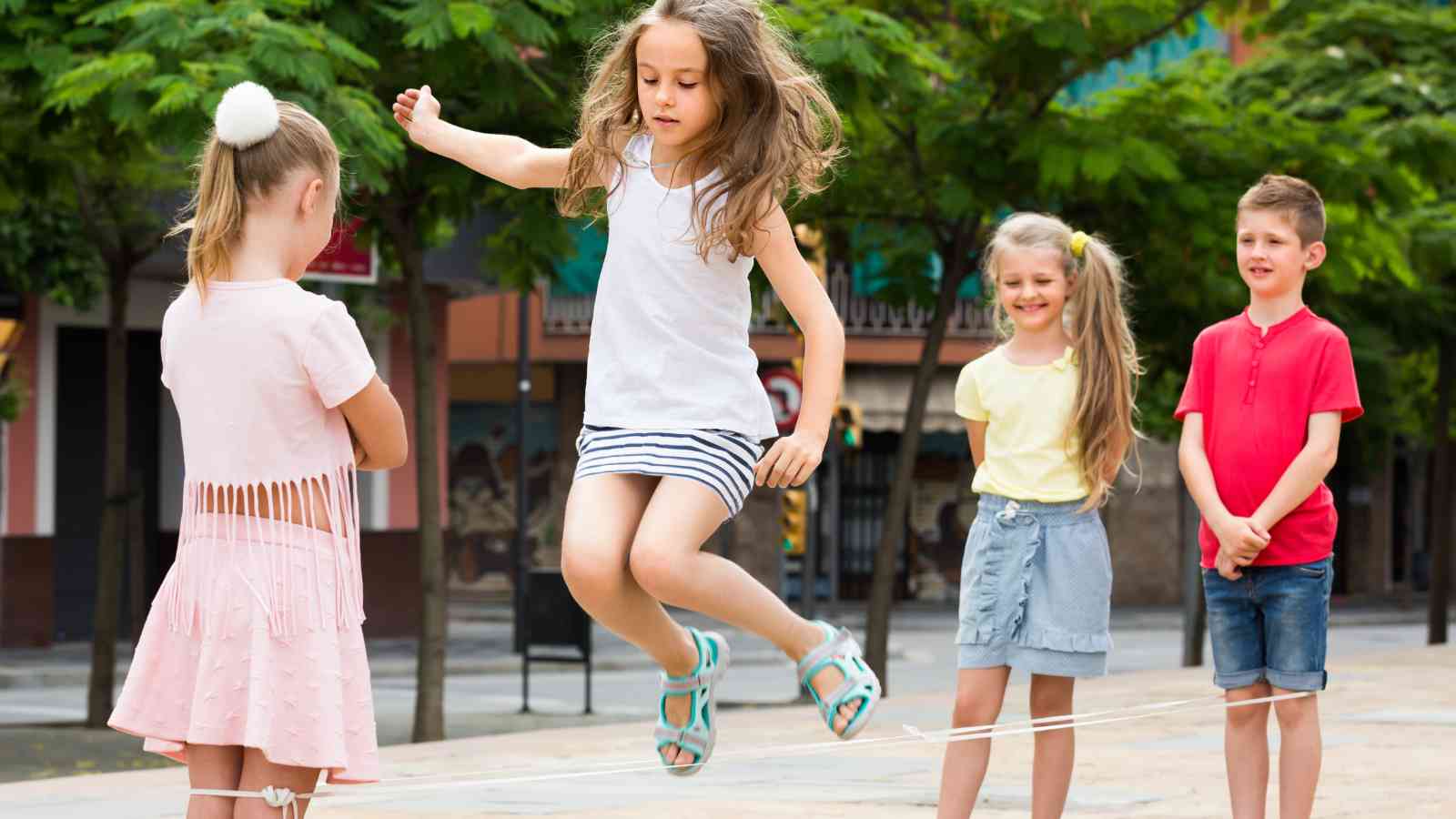 Four children play an elastic band jump game outdoors; one girl jumps while two girls hold the elastic and a boy stands nearby.