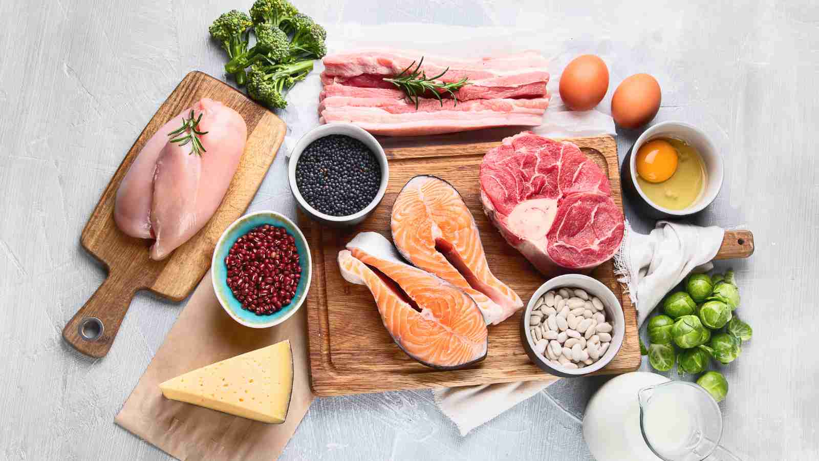 A variety of high-protein foods, including meat, fish, poultry, eggs, beans, and lentils, are arranged on a wooden cutting board and a light gray surface.