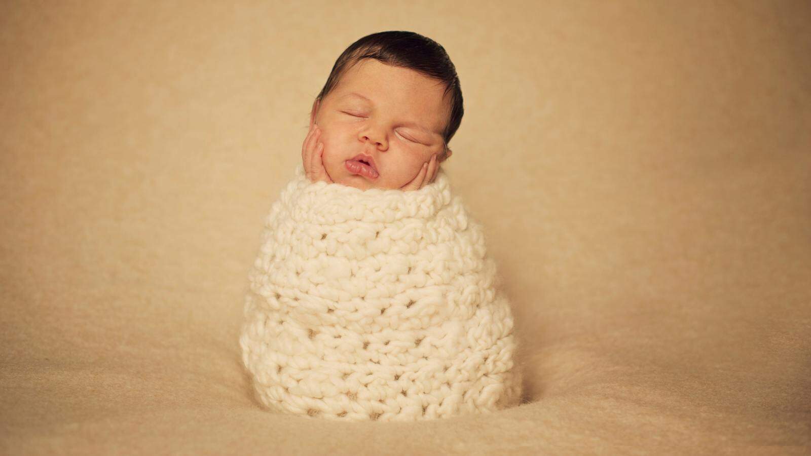 A newborn with dark hair is swaddled snugly in a thick, cream-colored knit blanket, sleeping upright with hands on their cheeks against a beige background.