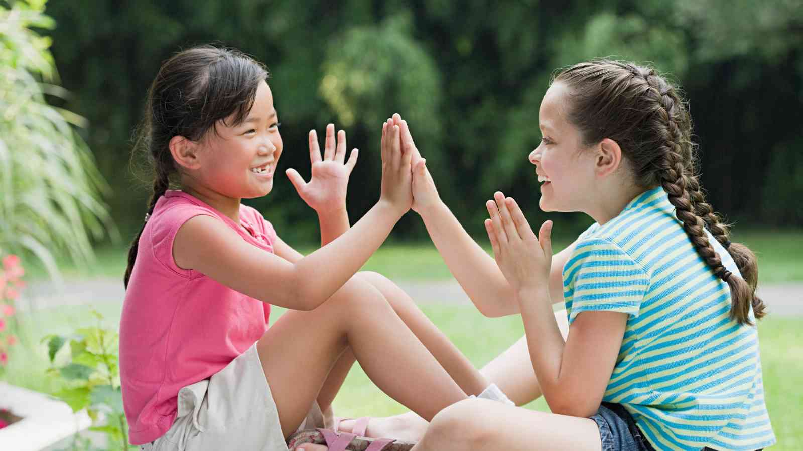 Two young girls sit facing each other outdoors, smiling and playing a hand-clapping game.