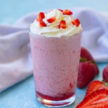 A glass of pink strawberry milkshake topped with whipped cream and chopped strawberries, with whole and sliced strawberries beside it on a blue surface.