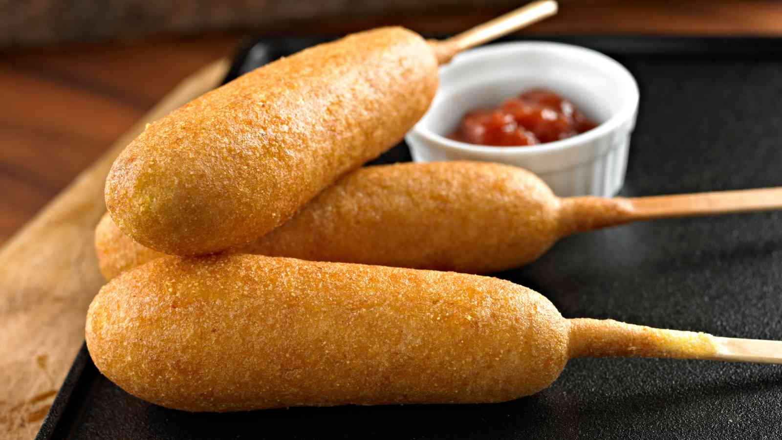 Three corn dogs on a black tray with a small cup of ketchup in the background.