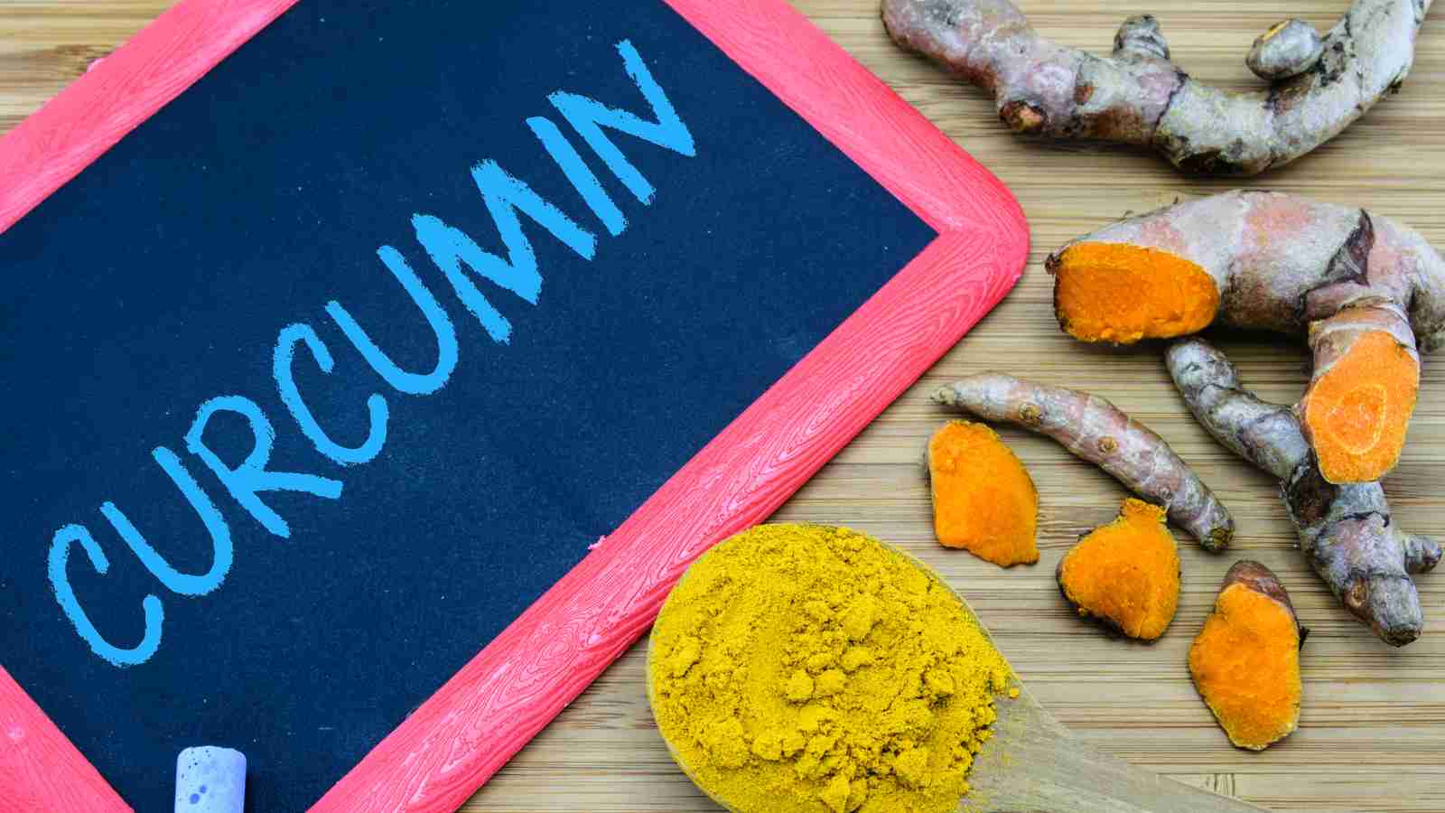 A small chalkboard with the word "Curcumin" written on it sits next to a wooden spoon of turmeric powder and several pieces of fresh turmeric root.