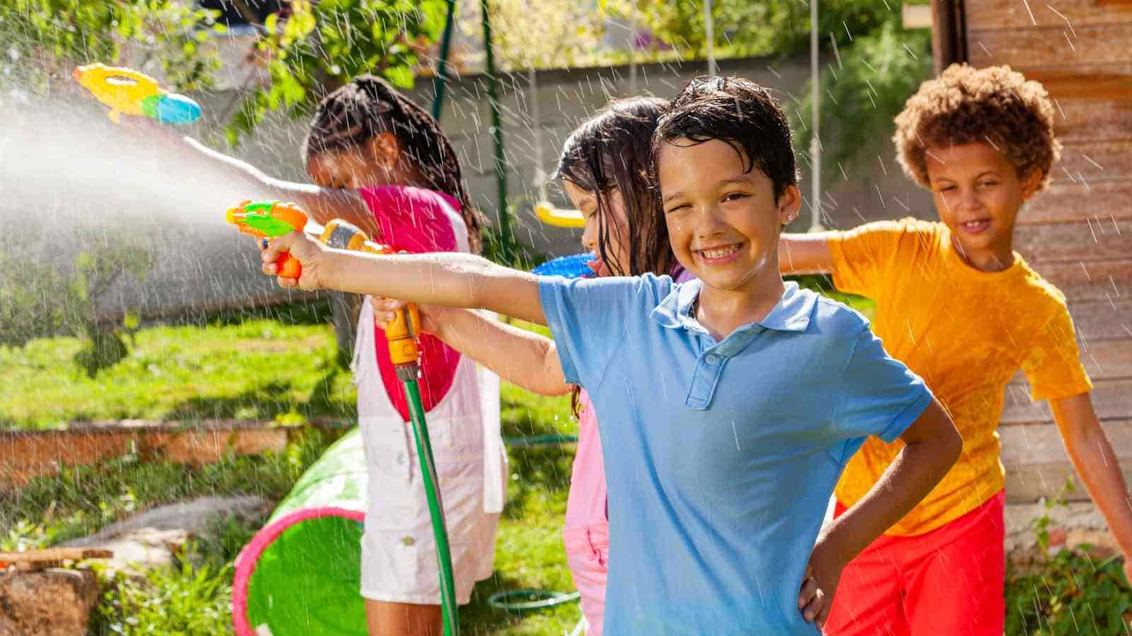 Four children play outside, smiling and spraying water with toy water guns on a sunny day, with grass and a playground structure in the background.