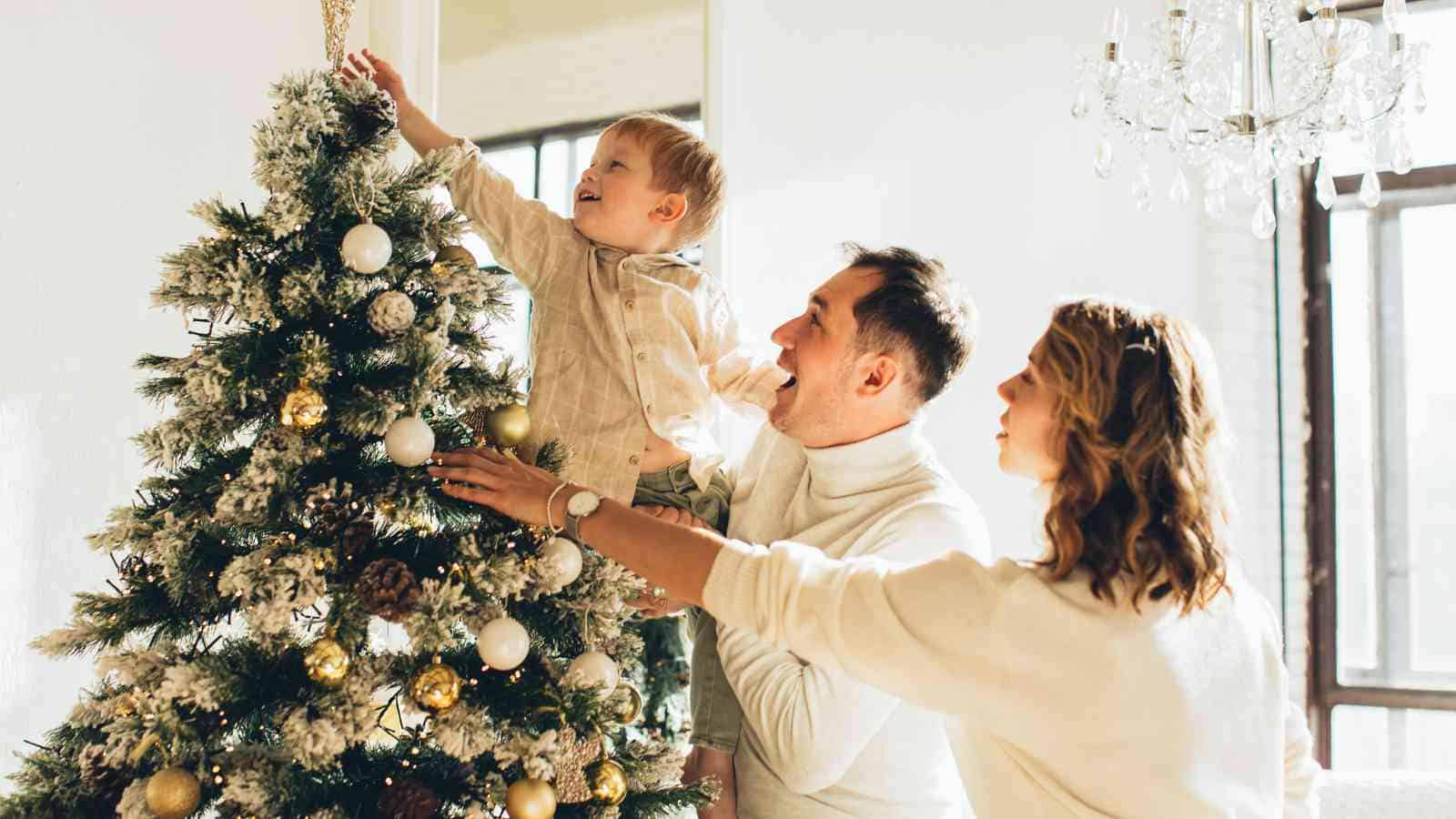 A family decorates a Christmas tree together; a child is reaching to place an ornament on a snowy, gold-and-white themed tree while two adults assist.