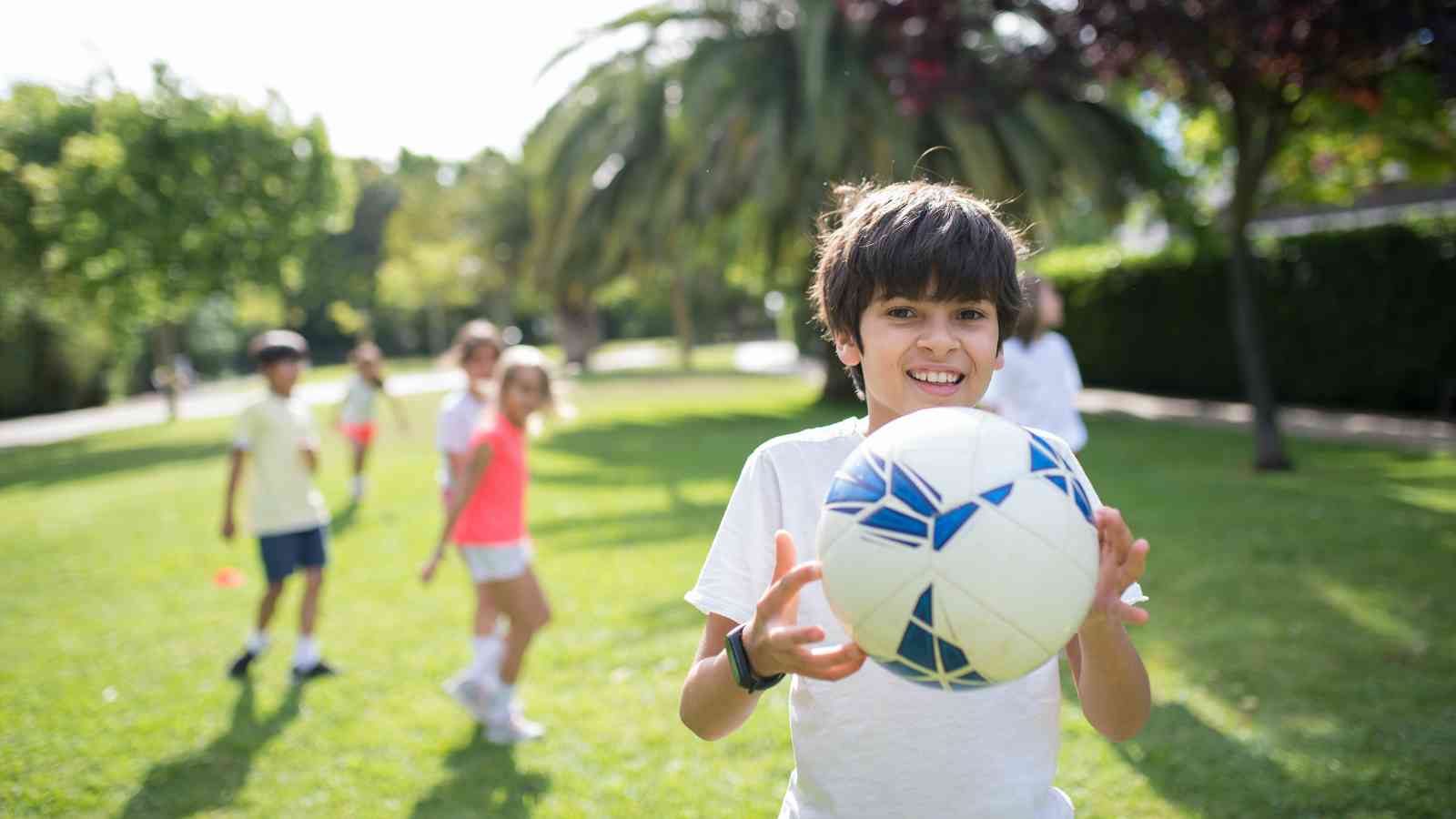 A boy holding a soccer ball stands on a grassy field, with other children and trees visible in the background on a sunny day.