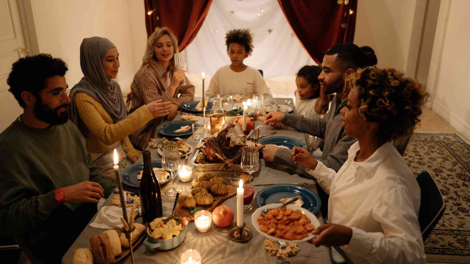 Seven people sit around a candlelit dinner table, sharing food and conversation in a warmly decorated room with curtains and string lights in the background.