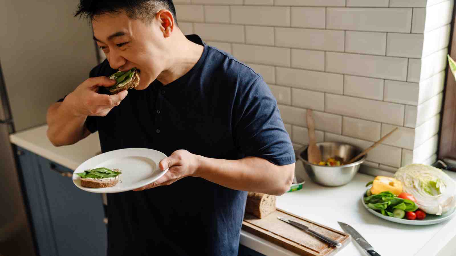 A man stands in a kitchen eating a slice of bread with greens, holding a plate, with fresh vegetables and a metal bowl on the counter beside him.