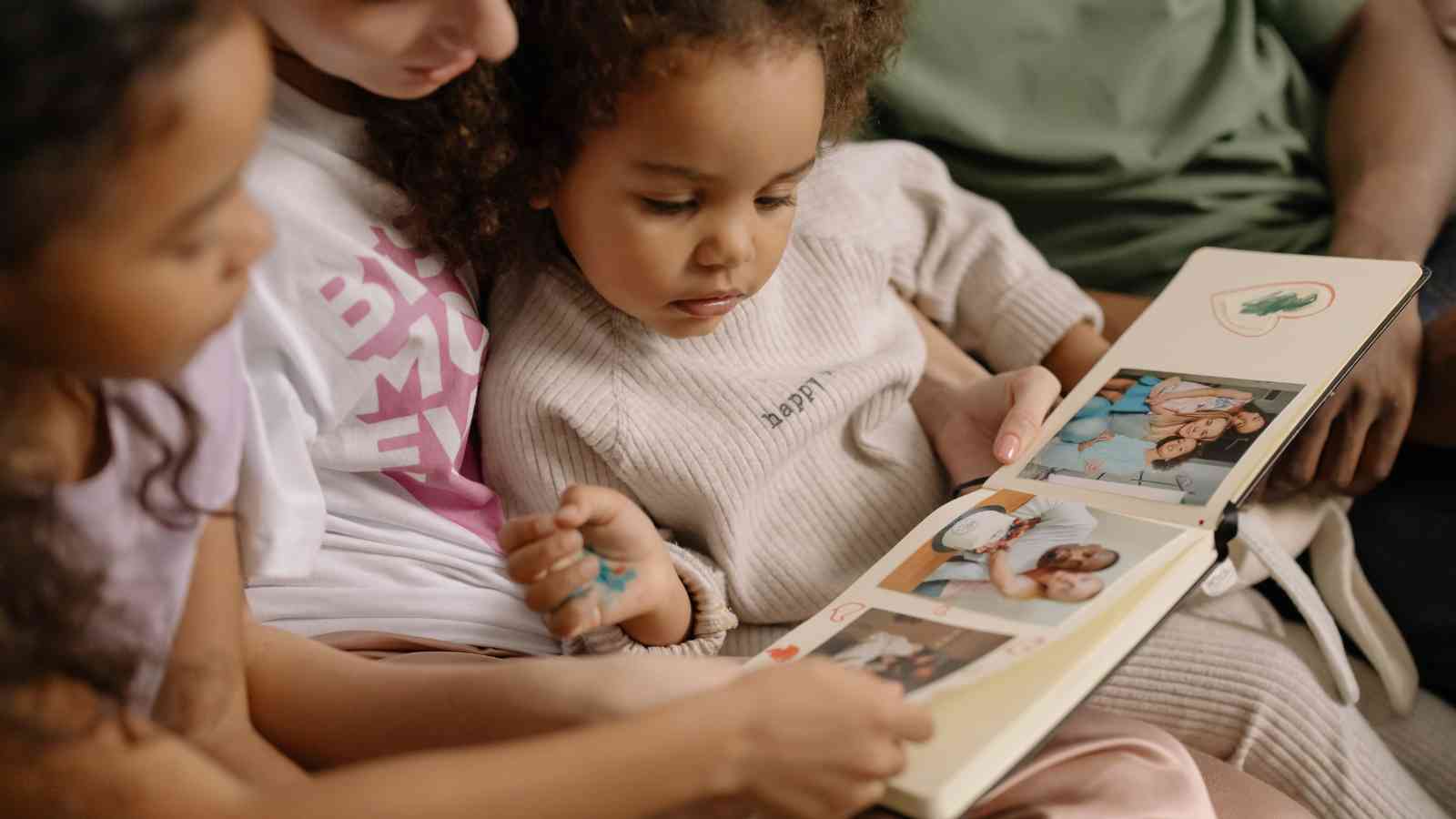 A child sits on an adult’s lap and looks at a photo album with family pictures, surrounded by other people.