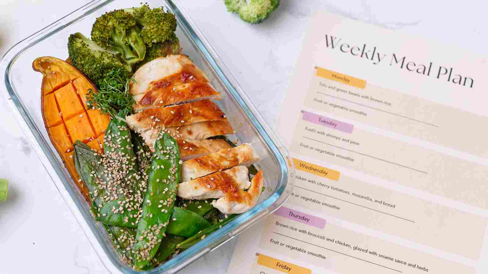 A glass container with grilled chicken, broccoli, snow peas with sesame seeds, and sweet potato next to a printed weekly meal plan.