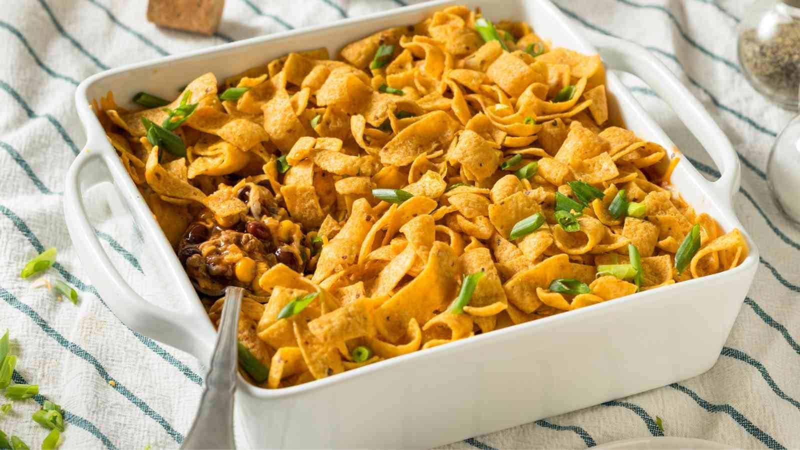 A white baking dish filled with Frito pie topped with corn chips and chopped green onions sits on a striped cloth. A serving spoon is visible in one corner.