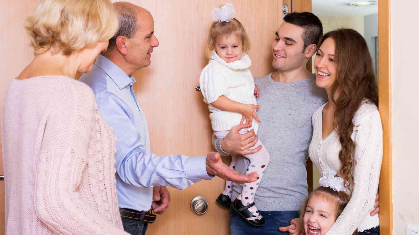 Two adults greet a family of four at the door; a man holds a young child, while a woman and another child stand beside him, all smiling.