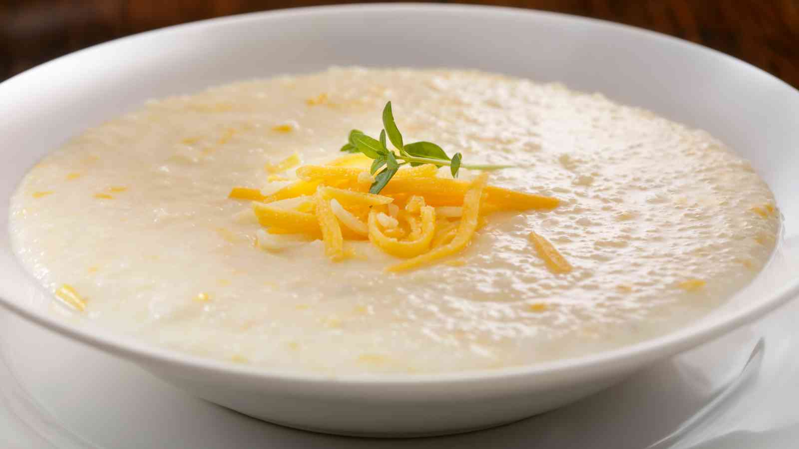A bowl of creamy grits topped with shredded cheese and a small sprig of fresh herbs, served in a white dish.