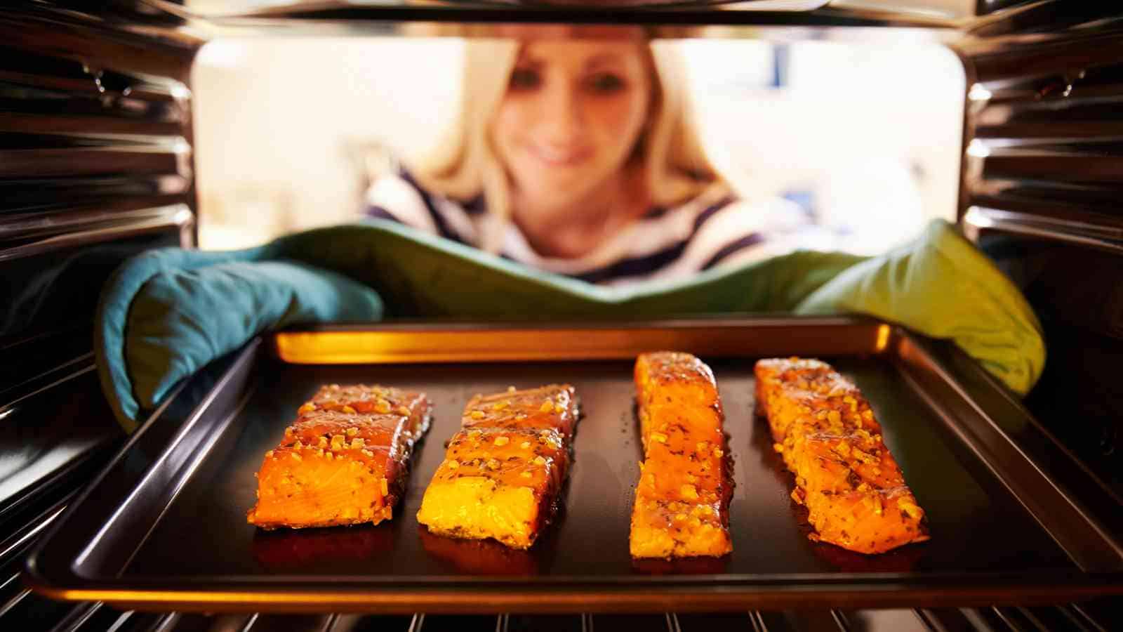 A woman wearing oven mitts places a baking tray with four seasoned salmon fillets into an oven.