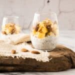 Two glasses filled with chia pudding, topped with diced pineapple, coconut flakes, and nuts, placed on a wooden board with a textured cloth and a few almonds.
