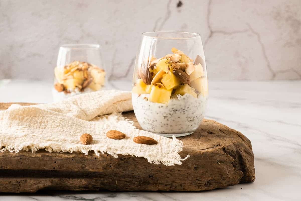 Two glasses filled with chia pudding, topped with diced pineapple, coconut flakes, and nuts, placed on a wooden board with a textured cloth and a few almonds.