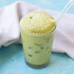 A glass of iced matcha latte with a frothy top and a clear straw, placed on a blue surface next to a light-colored cloth.