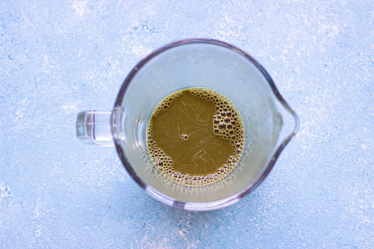A glass measuring cup containing a greenish-brown liquid, viewed from above, on a light blue textured surface.